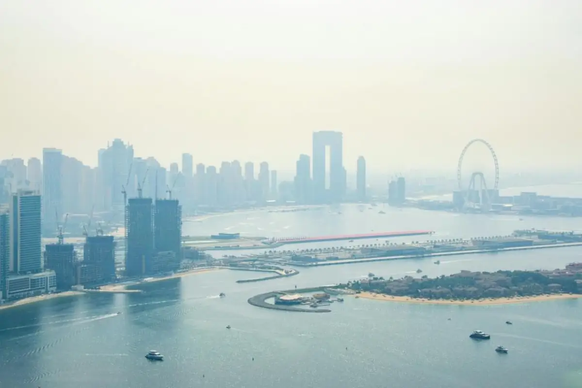Hazy aerial view of Dubai Marina's skyline and towers under construction, with Ain Dubai's observation wheel on Bluewaters Island and the edge of Palm Jumeirah across the water, boats crossing the bay below.